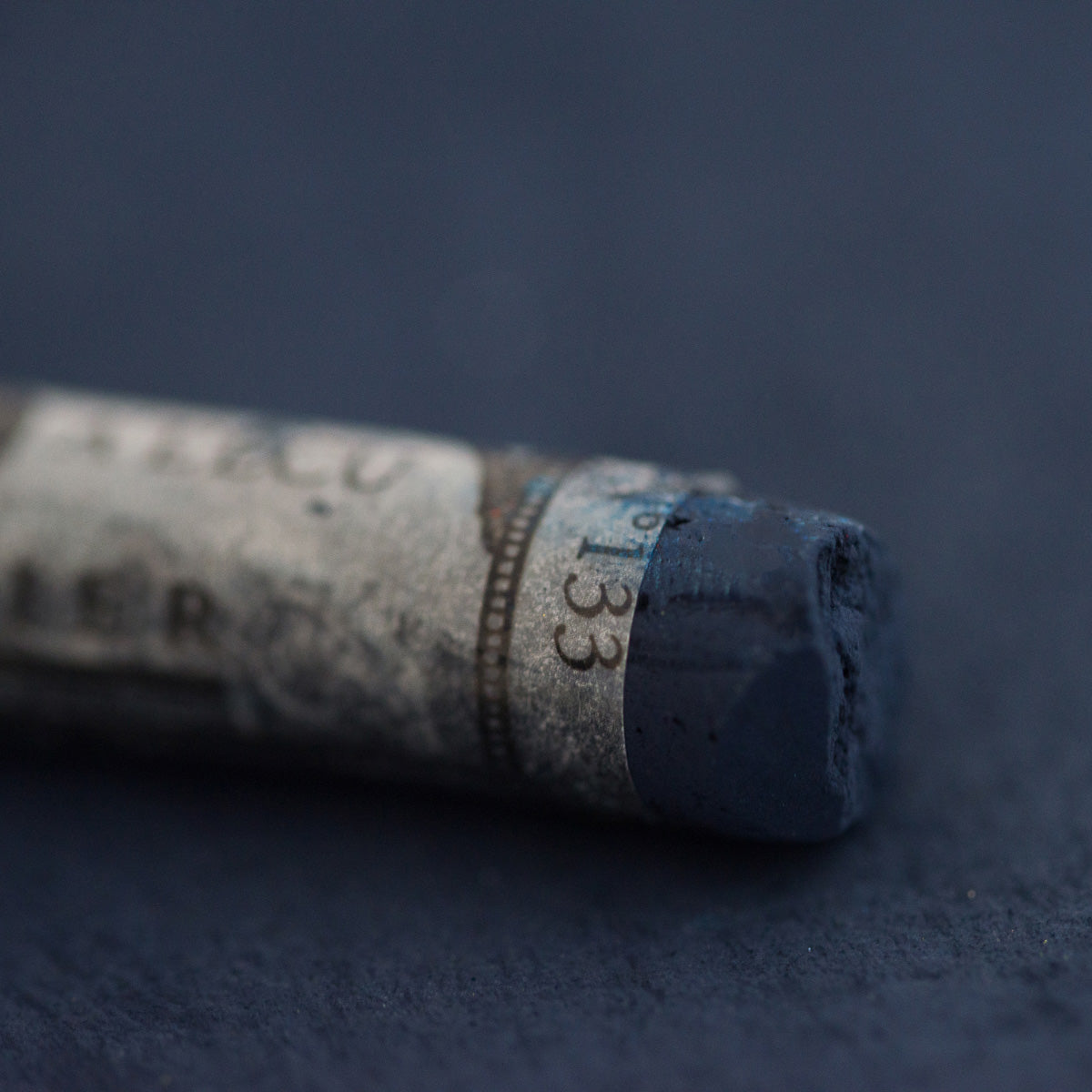 A close-up of a dark blue Sennelier extra soft pastel on the same coloured background, with the colour information printed on the paper wrap.
