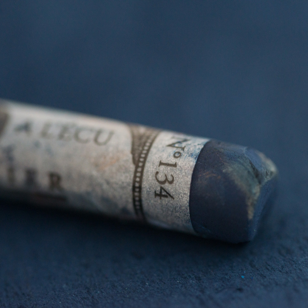A close-up of a blue Sennelier extra soft pastel on the same coloured background, with the colour information printed on the paper wrap.