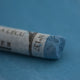 A close-up of a blue Sennelier extra soft pastel on the same coloured background, with the colour information printed on the paper wrap.