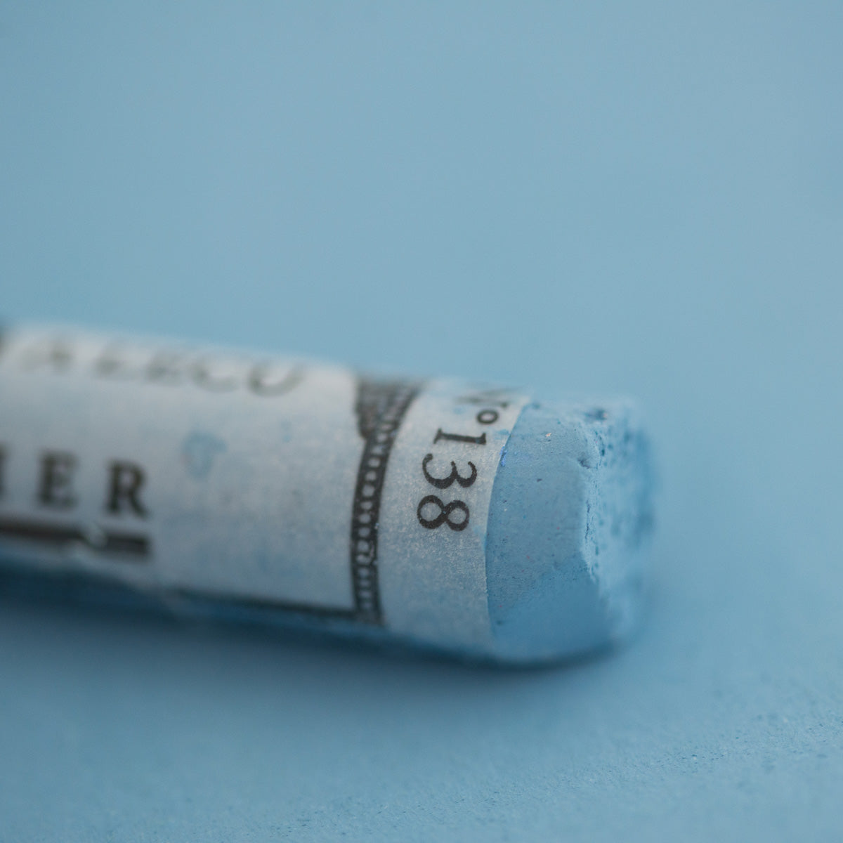 A close-up of a light blue Sennelier extra soft pastel on the same coloured background, with the colour information printed on the paper wrap.