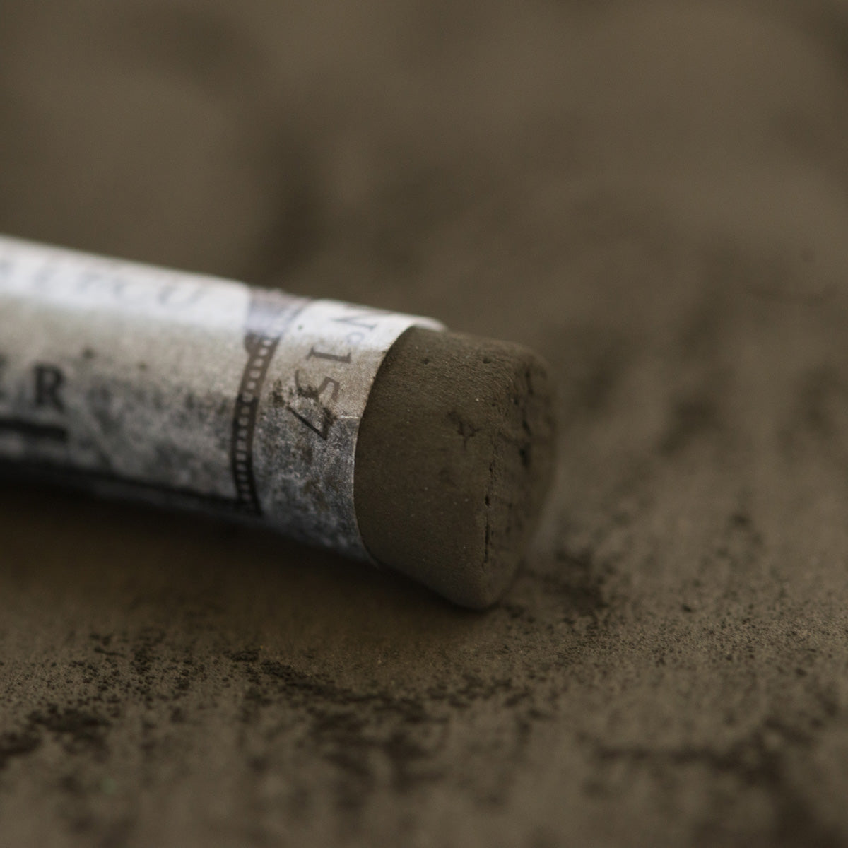 A close-up of a brown Sennelier extra soft pastel, on the same coloured background, with the colour information printed on the paper wrap.