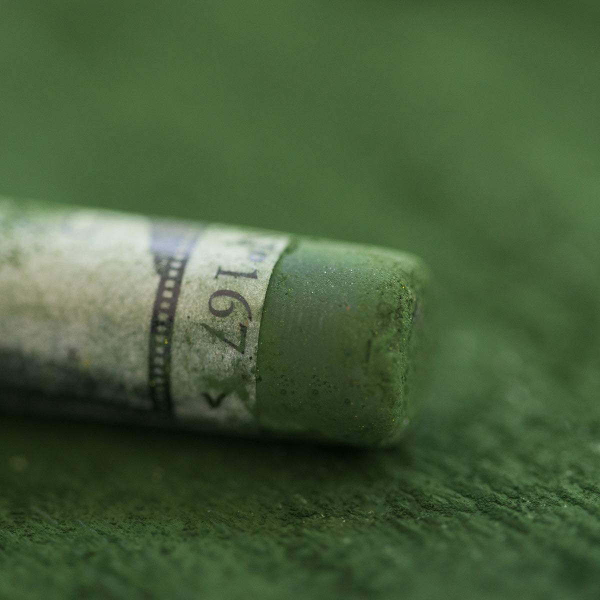 A close-up of a green Sennelier extra soft pastel. on the same coloured background, with the colour information printed on the paper wrap.