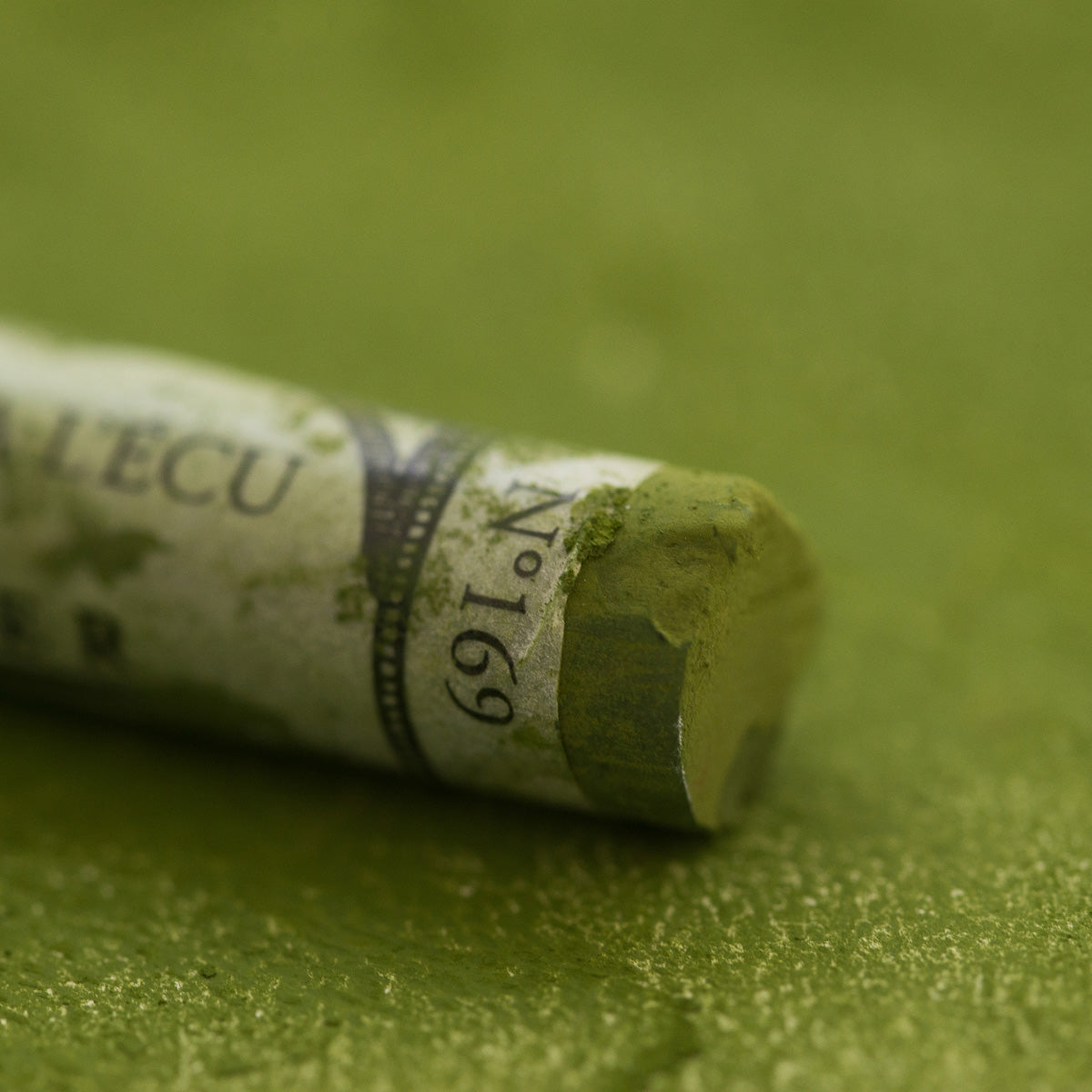 A close-up of a green Sennelier extra soft pastel. on the same coloured background, with the colour information printed on the paper wrap.