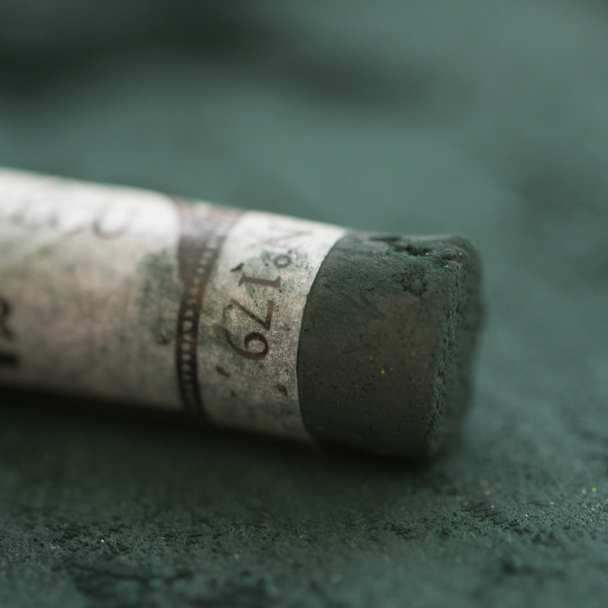 A close-up of a dark green Sennelier extra soft pastel, on the same coloured background, with the colour information printed on the paper wrap.