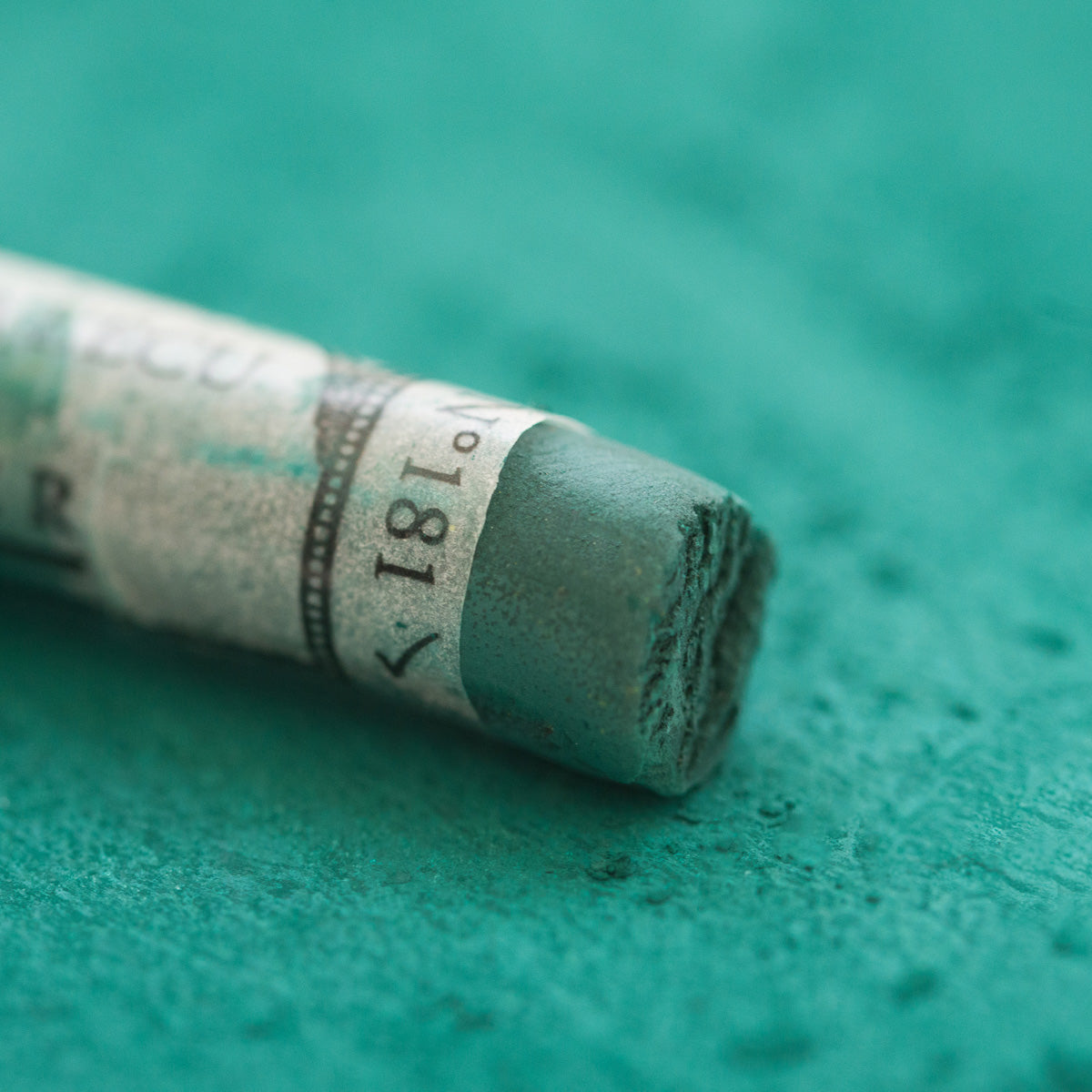 A close-up of a green Sennelier extra soft pastel, on the same coloured background, with the colour information printed on the paper wrap.