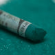 A close-up of a green Sennelier extra soft pastel, on the same coloured background, with the colour information printed on the paper wrap.