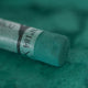 A close-up of a green Sennelier extra soft pastel, on the same coloured background, with the colour information printed on the paper wrap.
