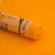 A close-up of an Orange Sennelier extra soft pastel. on the same coloured background, with the colour information printed on the paper wrap.