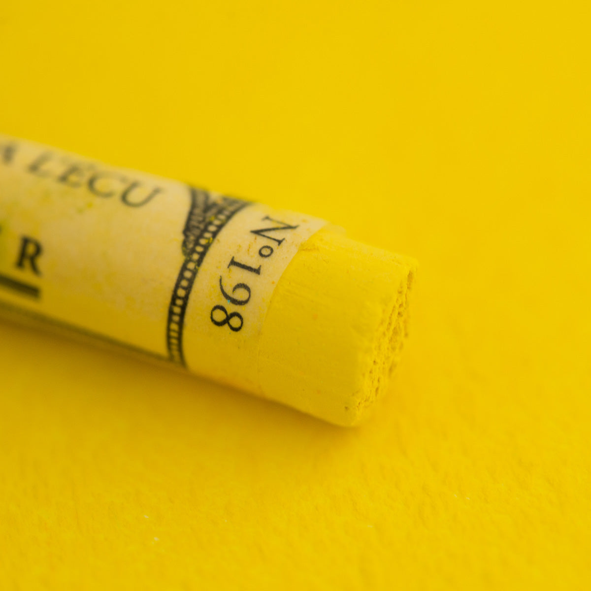 A close-up of a bright yellow Sennelier extra soft pastel. on the same coloured background, with the colour information printed on the paper wrap.