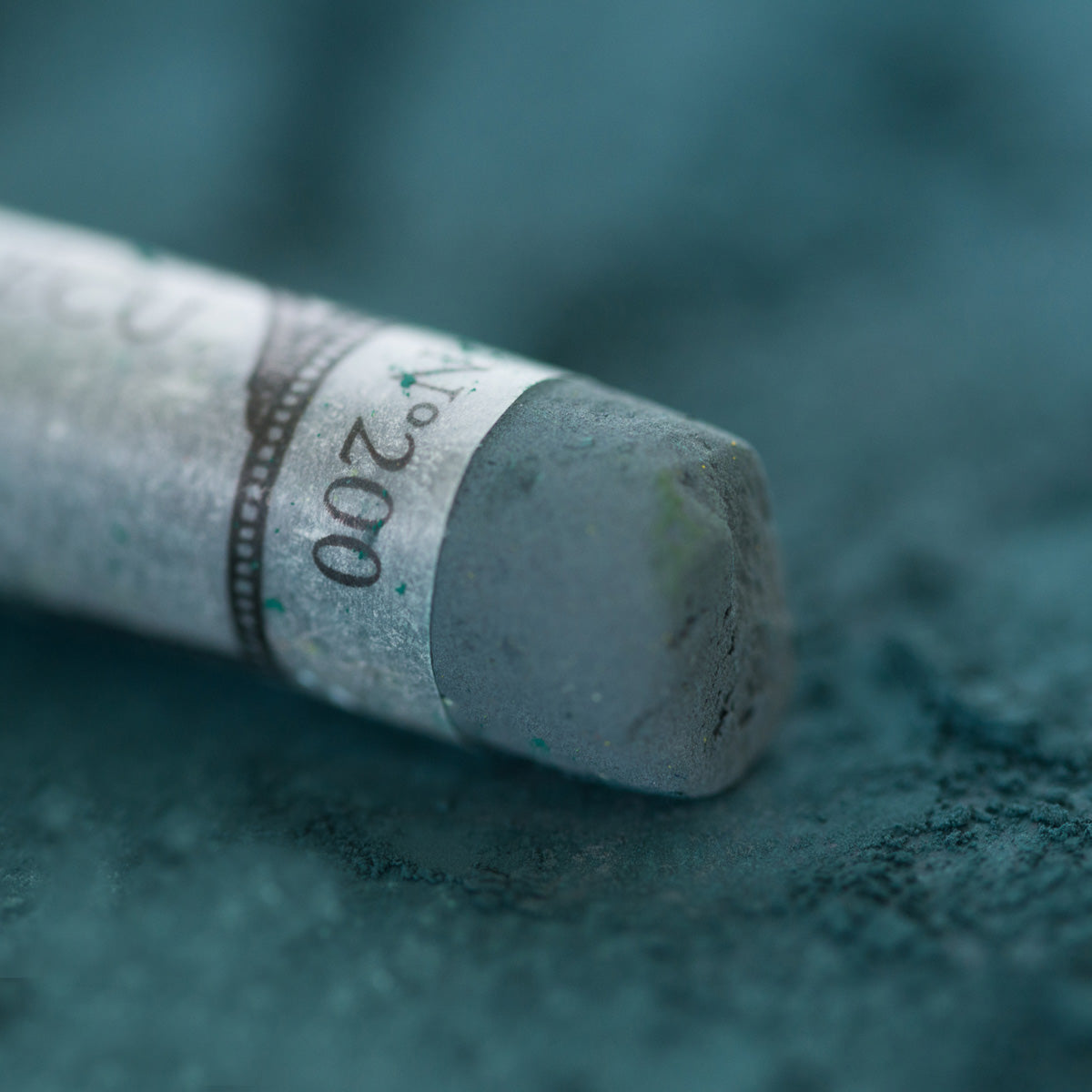 A close-up of a deep green Sennelier extra soft pastel, on the same coloured background, with the colour information printed on the paper wrap.