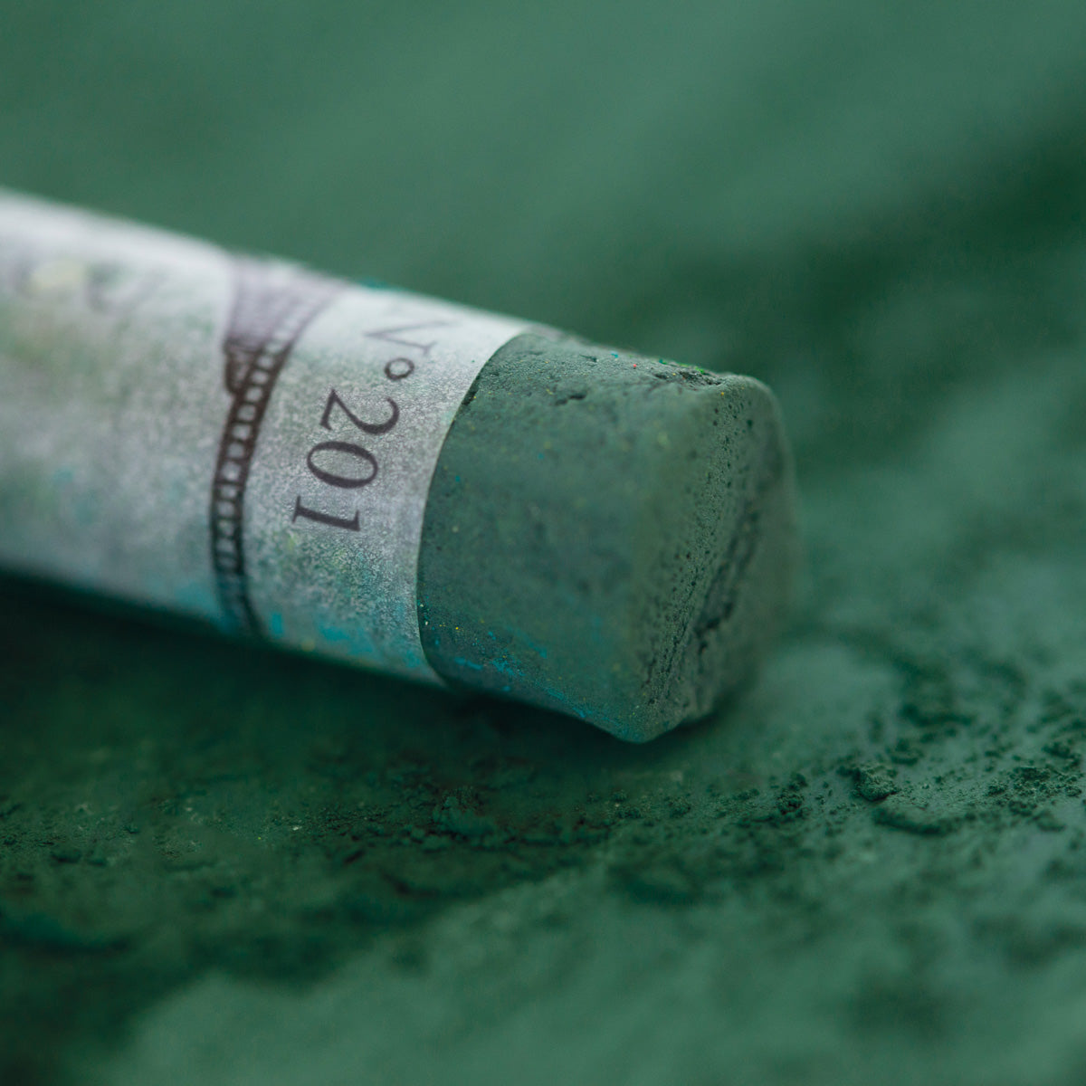A close-up of a green Sennelier extra soft pastel, on the same coloured background, with the colour information printed on the paper wrap.