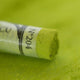 A close-up of a green Sennelier extra soft pastel, on the same coloured background, with the colour information printed on the paper wrap.