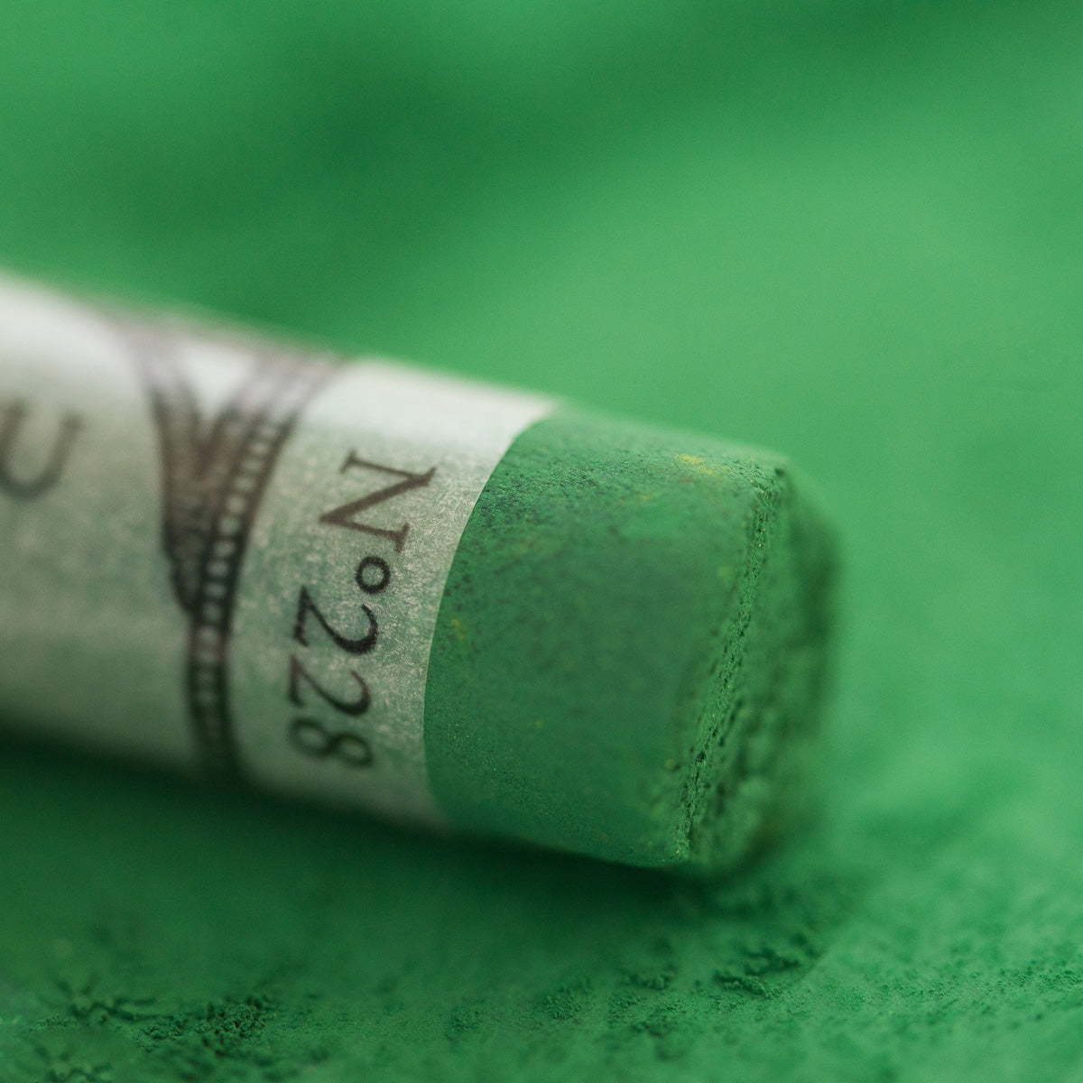 A close-up of a green Sennelier extra soft pastel, on the same coloured background, with the colour information printed on the paper wrap.