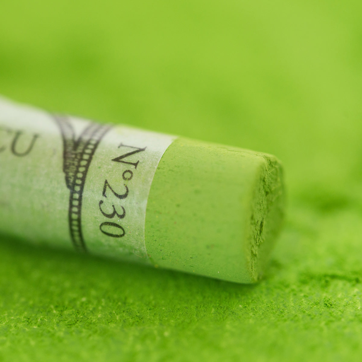 A close-up of a green Sennelier extra soft pastel, on the same coloured background, with the colour information printed on the paper wrap.