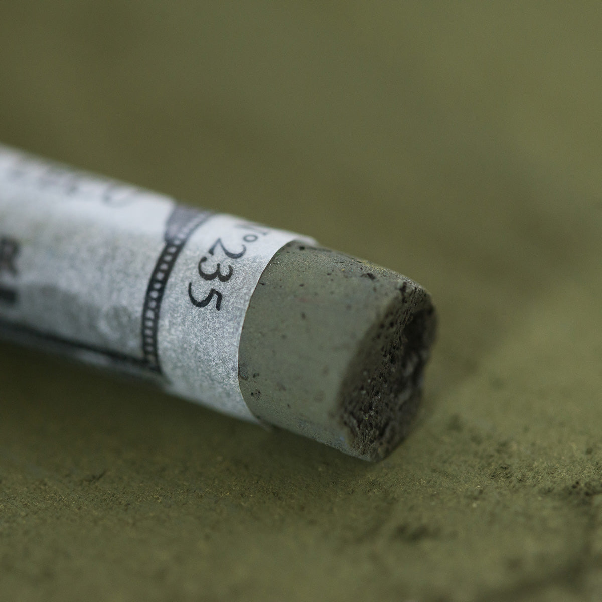 A close-up of a green Sennelier extra soft pastel, on the same coloured background, with the colour information printed on the paper wrap.