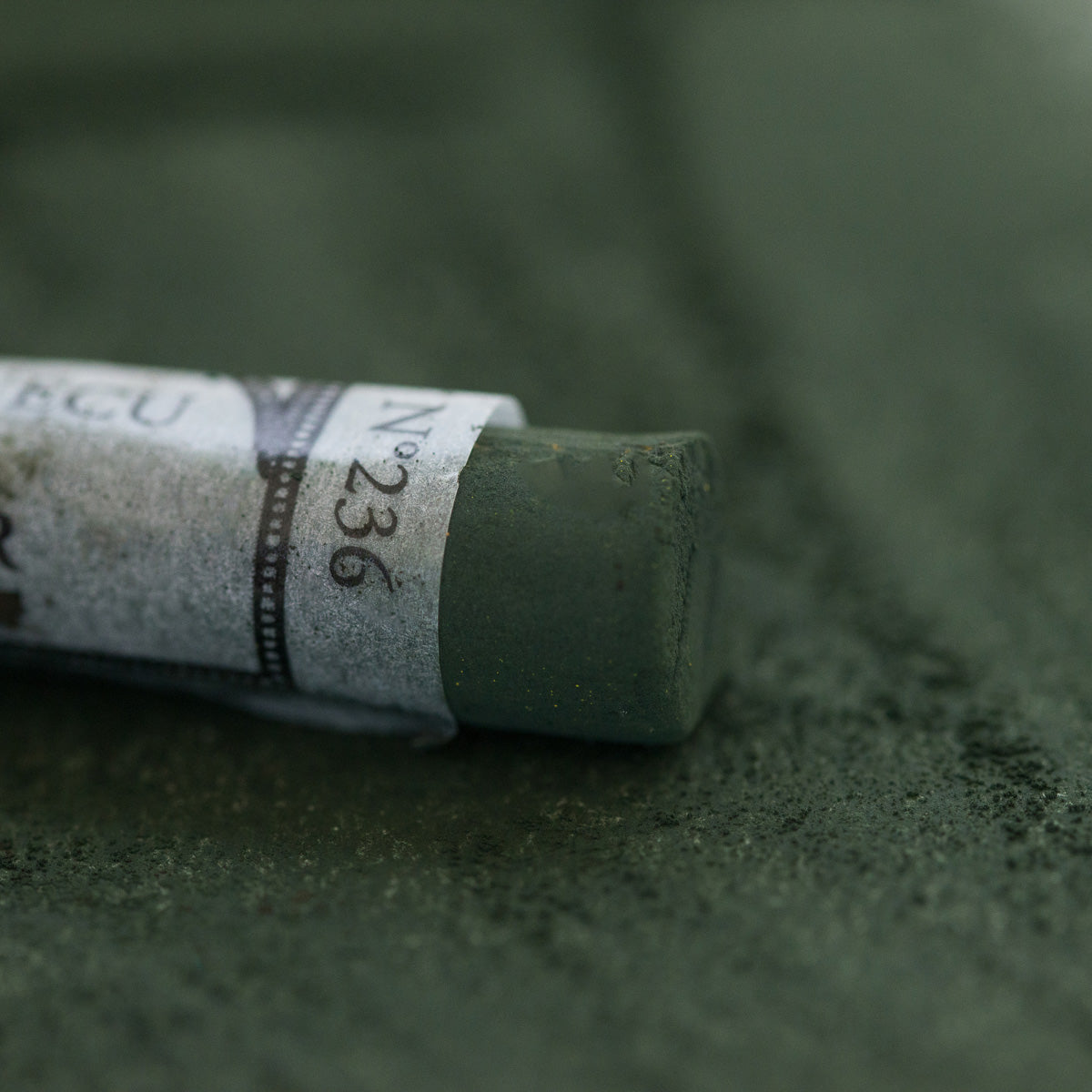 A close-up of a dark green Sennelier extra soft pastel, on the same coloured background, with the colour information printed on the paper wrap.