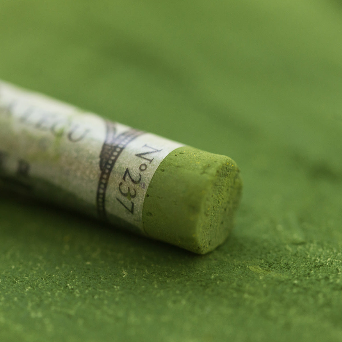 A close-up of a green Sennelier extra soft pastel, on the same coloured background, with the colour information printed on the paper wrap.