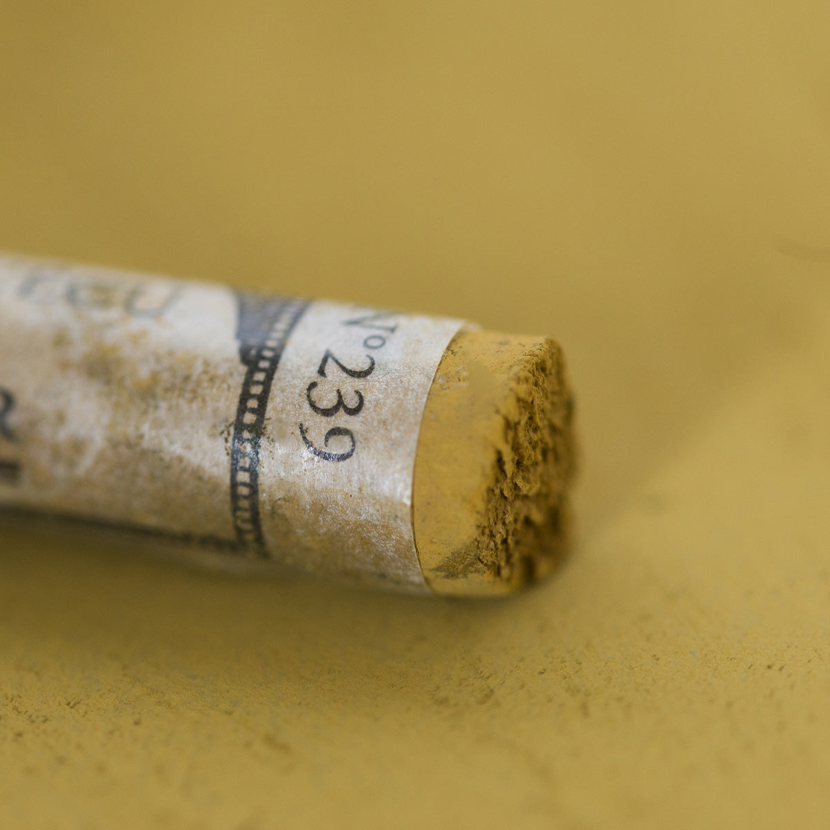 A close-up of a yellow-green Sennelier extra soft pastel, on the same coloured background, with the colour information printed on the paper wrap.