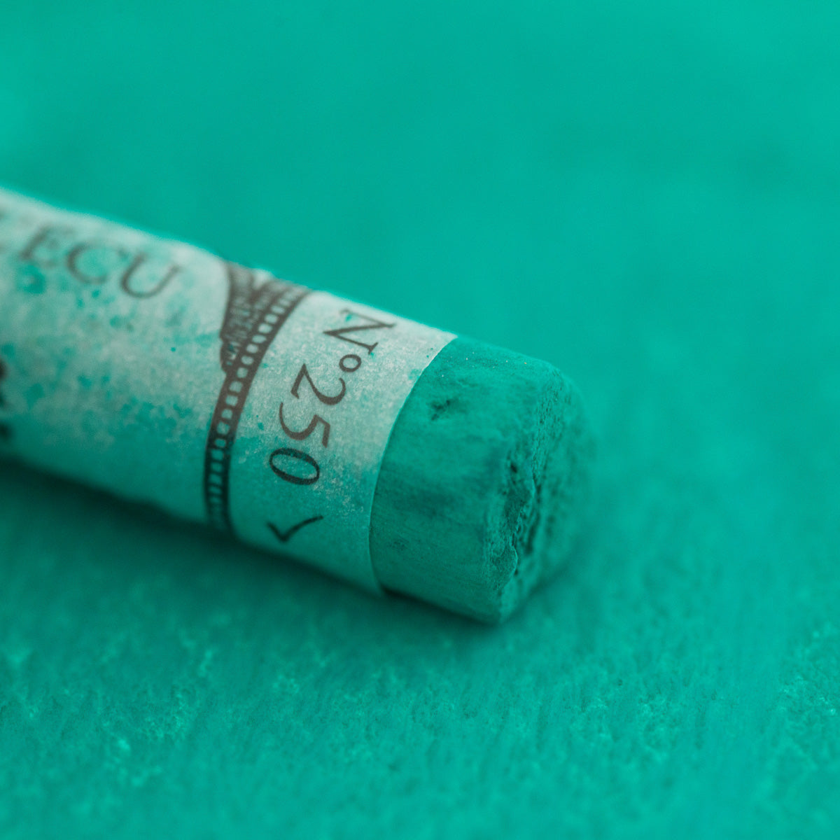 A close-up of a green Sennelier extra soft pastel. on the same coloured background, with the colour information printed on the paper wrap.