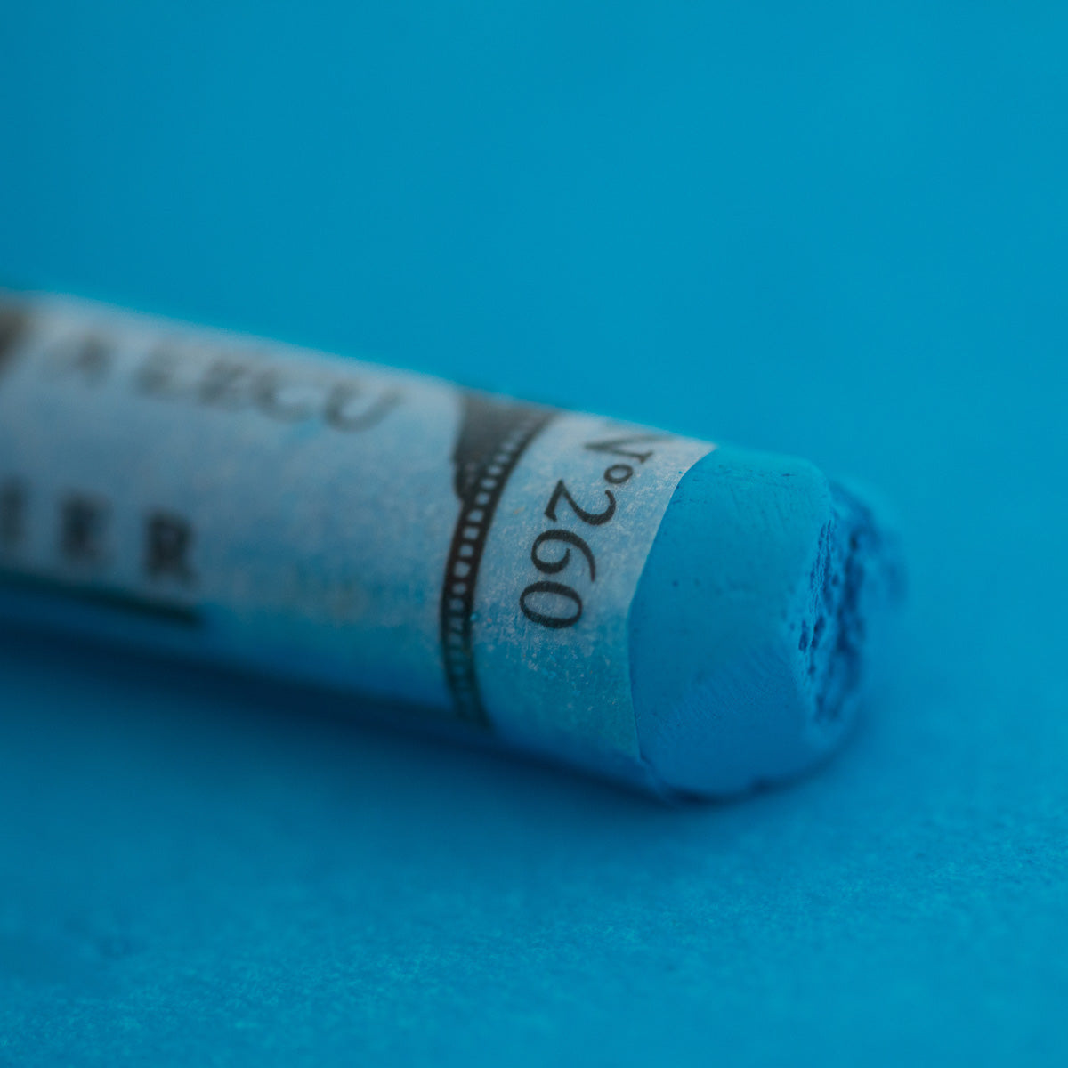 A close-up of a blue Sennelier extra soft pastel on the same coloured background, with the colour information printed on the paper wrap.