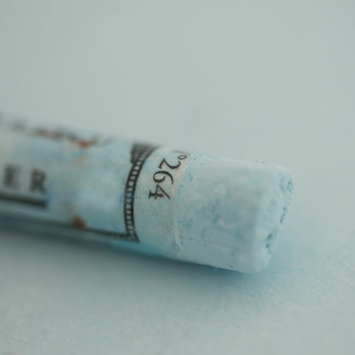 A close-up of a pale blue Sennelier extra soft pastel on the same coloured background, with the colour information printed on the paper wrap.