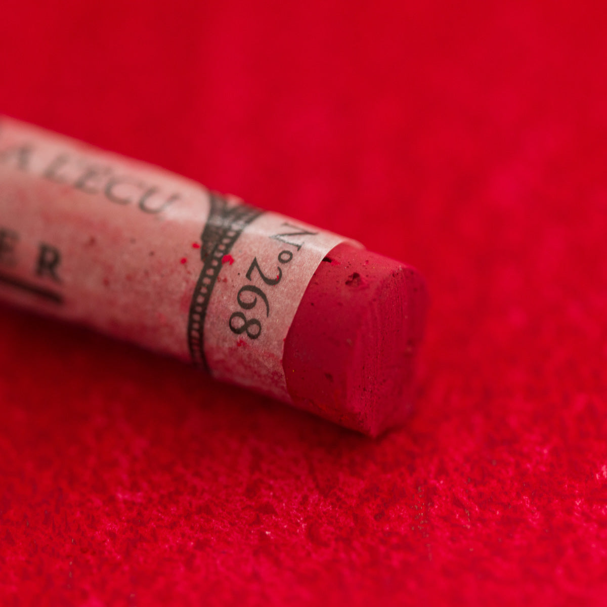 A close-up of a red Sennelier extra soft pastel on the same coloured background, with the colour information printed on the paper wrap.