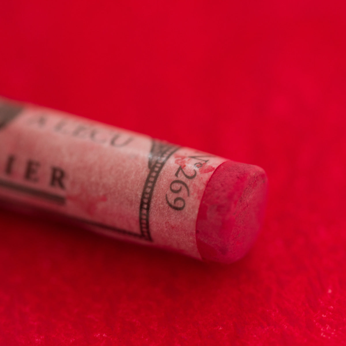 A close-up of a red Sennelier extra soft pastel on the same coloured background, with the colour information printed on the paper wrap.