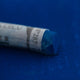 A close-up of a blue Sennelier extra soft pastel on the same coloured background, with the colour information printed on the paper wrap