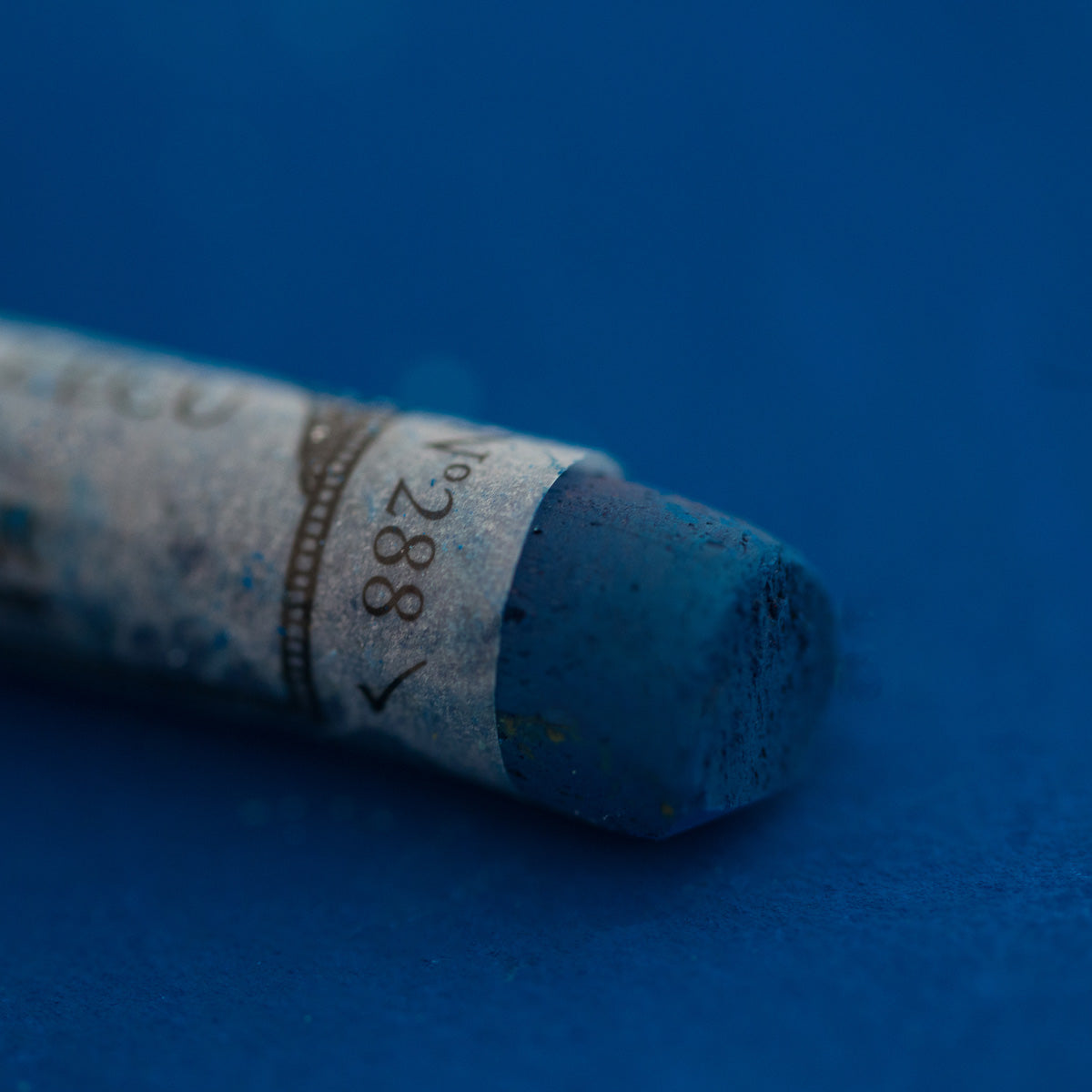 A close-up of a blue Sennelier extra soft pastel on the same coloured background, with the colour information printed on the paper wrap.