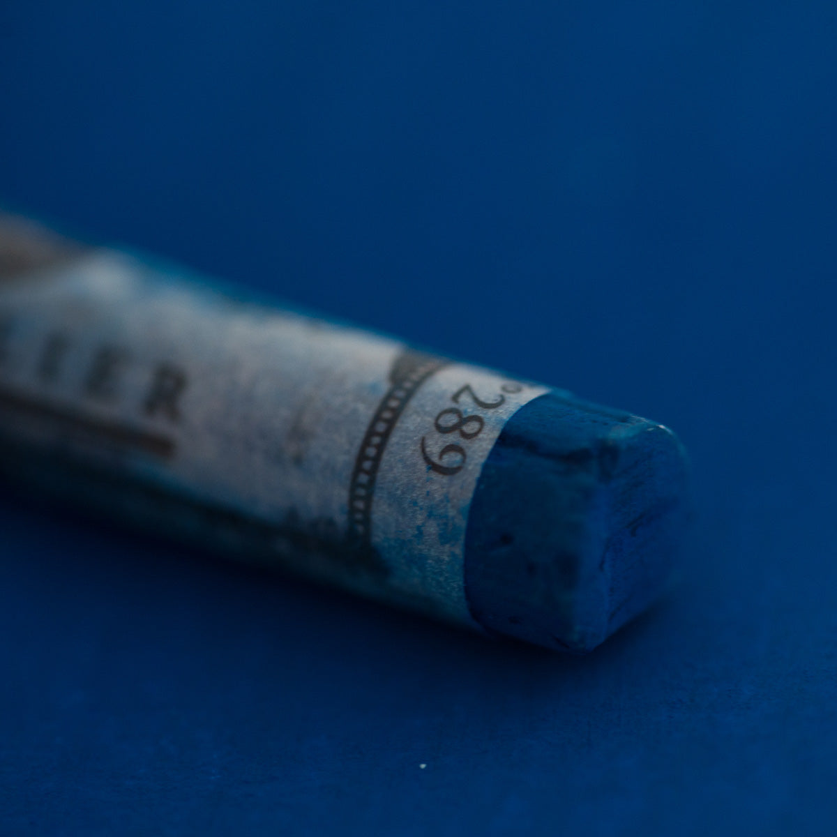 A close-up of a blue Sennelier extra soft pastel on the same coloured background, with the colour information printed on the paper wrap.