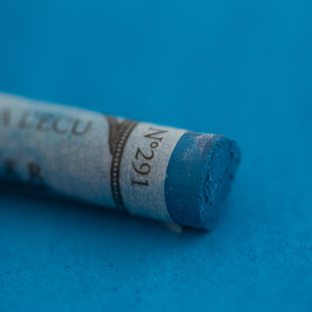 A close-up of a blue Sennelier extra soft pastel on the same coloured background, with the colour information printed on the paper wrap.