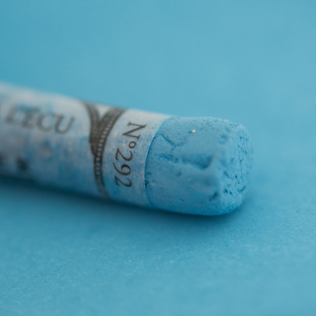 A close-up of a light blue Sennelier extra soft pastel on the same coloured background, with the colour information printed on the paper wrap.