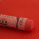 A close-up of a red Sennelier extra soft pastel on the same coloured background, with the colour information printed on the paper wrap.