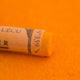 A close-up of a deep yellow Sennelier extra soft pastel. on the same coloured background, with the colour information printed on the paper wrap.