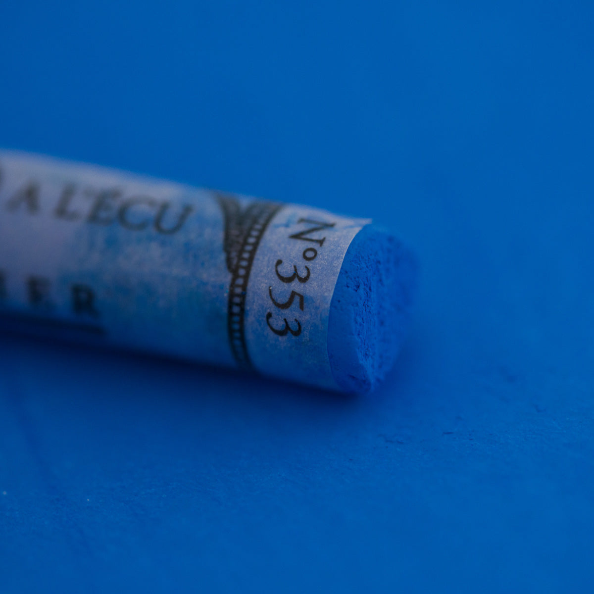 A close-up of a blue Sennelier extra soft pastel on the same coloured background, with the colour information printed on the paper wrap.
