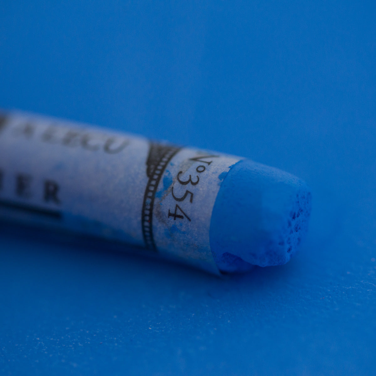 A close-up of a blue Sennelier extra soft pastel on the same coloured background, with the colour information printed on the paper wrap.