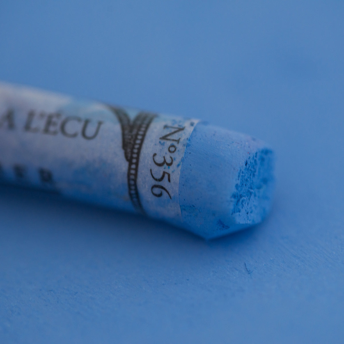 A close-up of a blue Sennelier extra soft pastel on the same coloured background, with the colour information printed on the paper wrap.