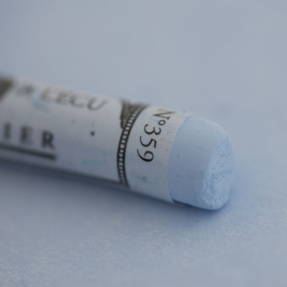 A close-up of a light blue Sennelier extra soft pastel on the same coloured background, with the colour information printed on the paper wrap.