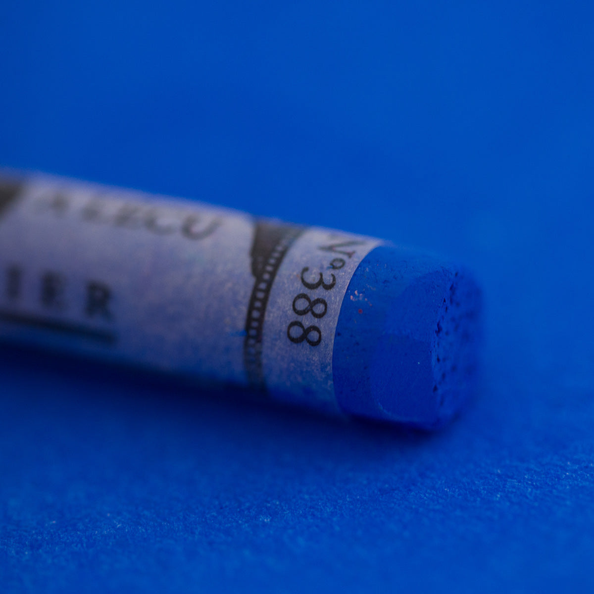 A close-up of a bright blue Sennelier extra soft pastel on the same coloured background, with the colour information printed on the paper wrap.