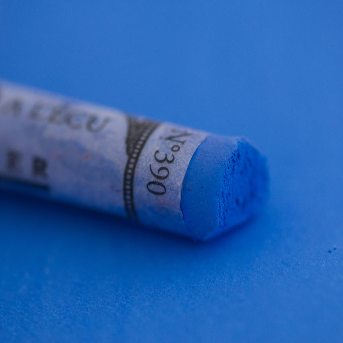 A close-up of a blue Sennelier extra soft pastel on the same coloured background, with the colour information printed on the paper wrap.