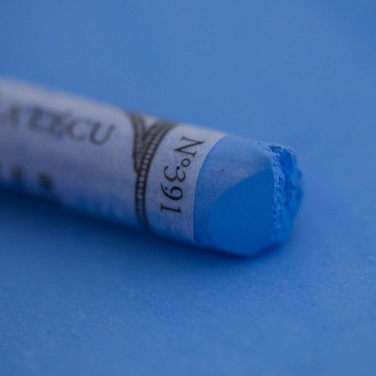 A close-up of a blue Sennelier extra soft pastel on the same coloured background, with the colour information printed on the paper wrap.