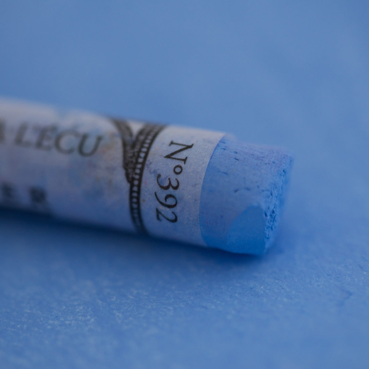 A close-up of a blue Sennelier extra soft pastel on the same coloured background, with the colour information printed on the paper wrap.