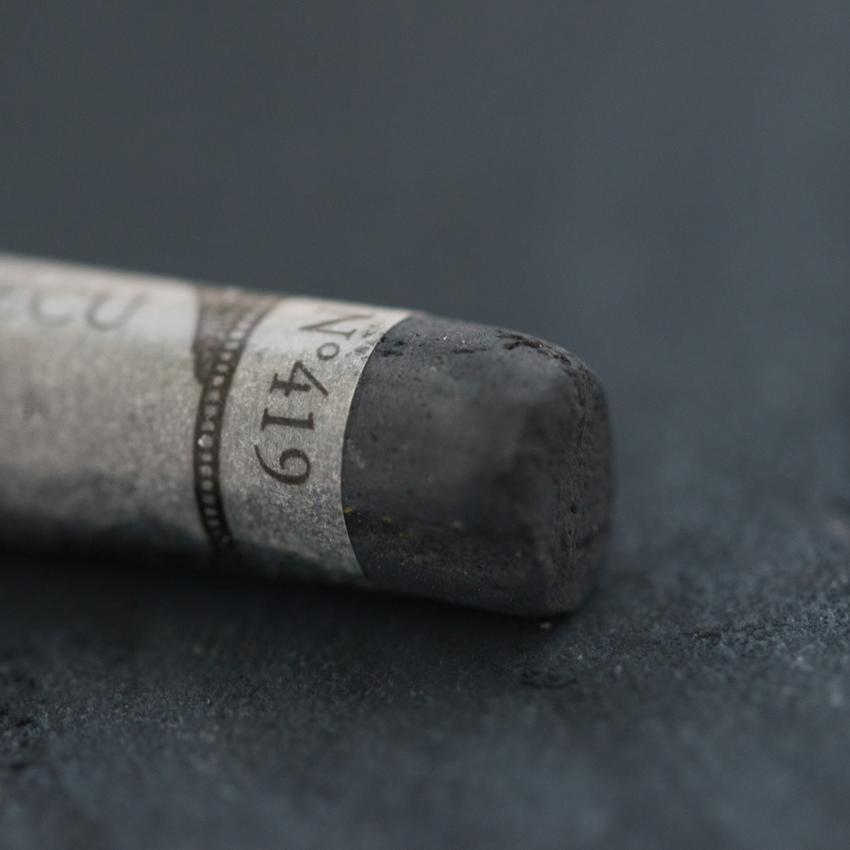 A close-up of a dark grey Sennelier extra soft pastel. on the same coloured background, with the colour information printed on the paper wrap.