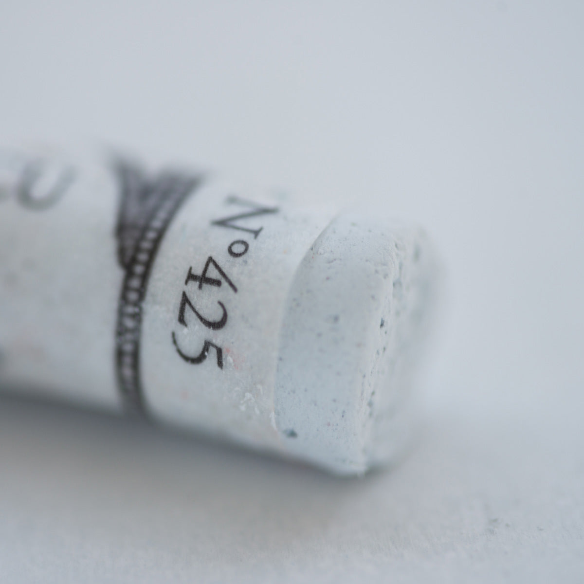 A close-up of a white Sennelier extra soft pastel. on the same coloured background, with the colour information printed on the paper wrap.