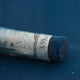 A close-up of a blue Sennelier extra soft pastel on the same coloured background, with the colour information printed on the paper wrap.