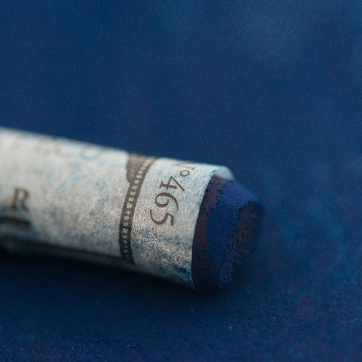 A close-up of a blue Sennelier extra soft pastel on the same coloured background, with the colour information printed on the paper wrap.