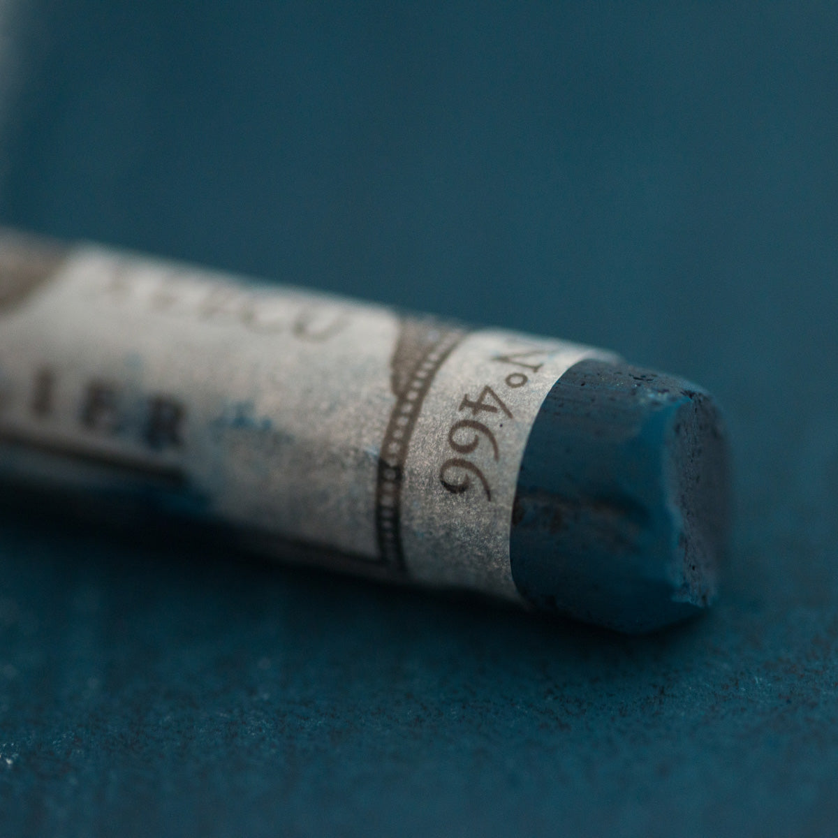 A close-up of a blue Sennelier extra soft pastel on the same coloured background, with the colour information printed on the paper wrap.