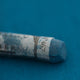 A close-up of a blue Sennelier extra soft pastel on the same coloured background, with the colour information printed on the paper wrap.