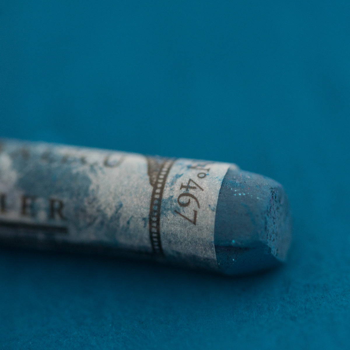 A close-up of a blue Sennelier extra soft pastel on the same coloured background, with the colour information printed on the paper wrap.
