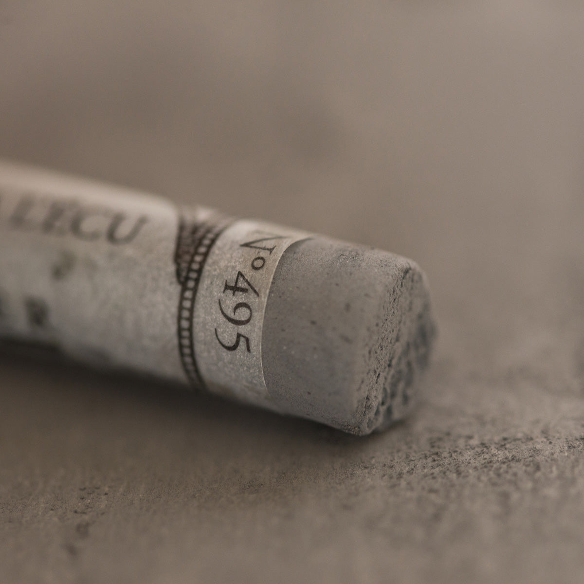 A close-up of a grey Sennelier extra soft pastel. on the same coloured background, with the colour information printed on the paper wrap.